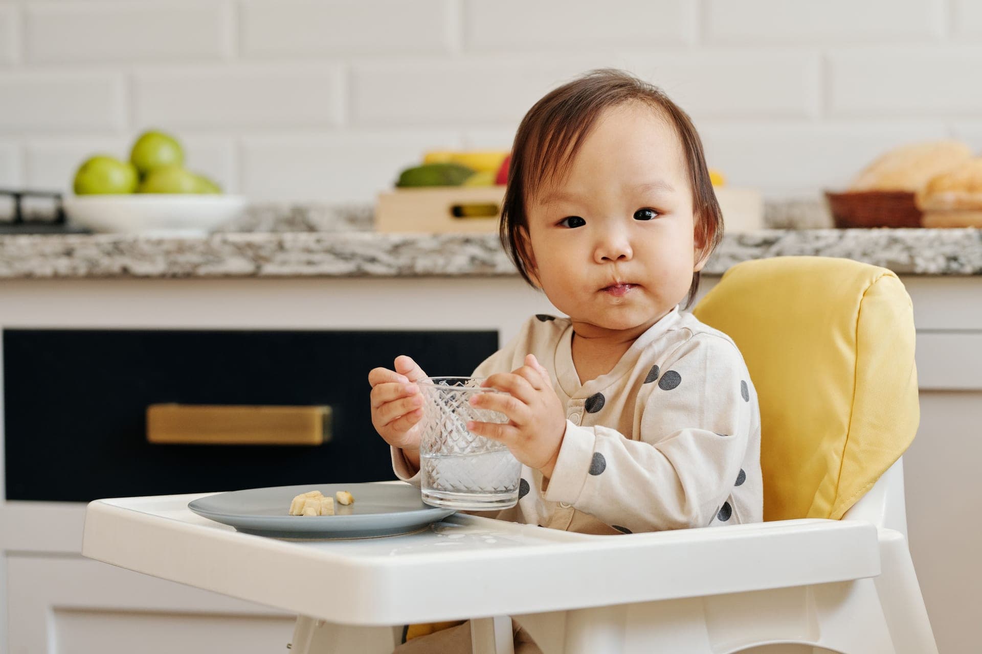 A toddler in a high chair enjoying a meal and a drink in a kitchen (illustrative photo)